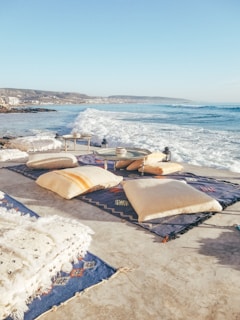 A coastal scene with large waves crashing onto a beachside patio or terrace. The area is decorated with colorful blankets and pillows arranged on the ground. Small tables with decorative items are placed on the rugs. In the distance, a coastline with buildings and hills can be seen under a clear blue sky.