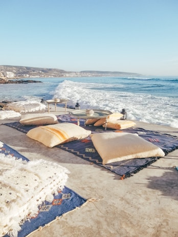 A coastal scene with large waves crashing onto a beachside patio or terrace. The area is decorated with colorful blankets and pillows arranged on the ground. Small tables with decorative items are placed on the rugs. In the distance, a coastline with buildings and hills can be seen under a clear blue sky.