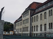 An old, imposing building with a weathered facade featuring numerous barred windows and a red-tiled roof. The structure suggests a historical or institutional architecture. In the foreground, there is a portion of a sports field with a goal post. Barbed wire is visible on a wall, suggesting security measures, and trees with dense foliage are present in the background.