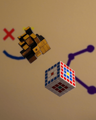 Two three-dimensional puzzles hover in midair. On the left is a complex wooden puzzle cube with interlocking pieces. On the right is a standard 4x4x4 Rubik's cube with various colored stickers. The background features simple graphical elements including a red X and blue and purple lines creating a casual and playful ambiance.