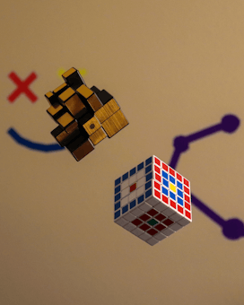 Two three-dimensional puzzles hover in midair. On the left is a complex wooden puzzle cube with interlocking pieces. On the right is a standard 4x4x4 Rubik's cube with various colored stickers. The background features simple graphical elements including a red X and blue and purple lines creating a casual and playful ambiance.