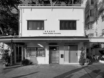 A two-story building with a simple architectural design. The upper level features three windows with shutters, and there's a sign reading 'Stanley Residents Association'. The lower level has a door with a wooden appearance, windows with metal security grilles, and potted plants on either side. The structure appears to be in an urban setting, possibly surrounded by residential buildings and greenery.