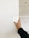A hand holding up a color swatch against a white wall in a room under construction. The wall is partially painted, and there is a visible hole in the ceiling.