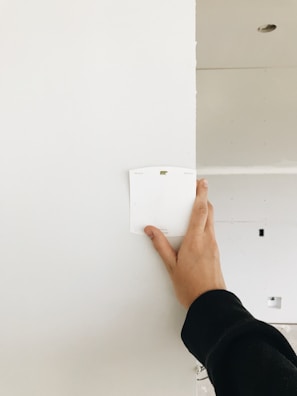 A hand holding up a color swatch against a white wall in a room under construction. The wall is partially painted, and there is a visible hole in the ceiling.