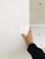 A hand holding up a color swatch against a white wall in a room under construction. The wall is partially painted, and there is a visible hole in the ceiling.