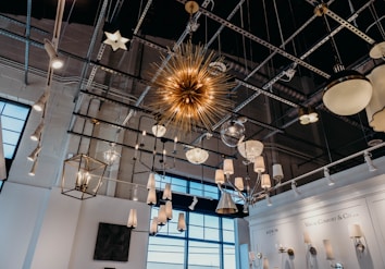 An array of diverse hanging light fixtures displayed in a showroom. The ceiling features a metallic grid with a variety of chandeliers, pendant lights, and spotlights. A prominent spiky golden chandelier is central. The walls are lined with sconce displays under a company branding plaque. Natural light filters in through large windows, enhancing the modern industrial feel.