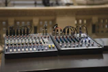 Two audio mixing consoles with numerous knobs, sliders, and connected cables placed on a dark surface. The background is blurred, featuring some architectural elements, possibly a balcony or railing with small rectangular openings.