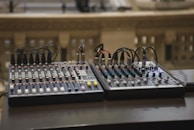 Two audio mixing consoles with numerous knobs, sliders, and connected cables placed on a dark surface. The background is blurred, featuring some architectural elements, possibly a balcony or railing with small rectangular openings.