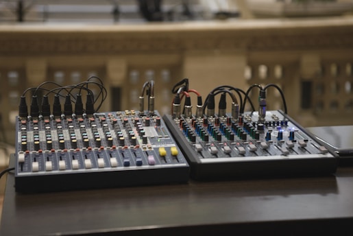 Two audio mixing consoles with numerous knobs, sliders, and connected cables placed on a dark surface. The background is blurred, featuring some architectural elements, possibly a balcony or railing with small rectangular openings.