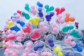 A large cluster of transparent balloons filled with colorful Mickey Mouse-shaped balloons, including pink, blue, green, red, and yellow hues, is held by someone in an outdoor amusement park setting. The background includes a castle and various people walking around, enhancing the lively and playful atmosphere.