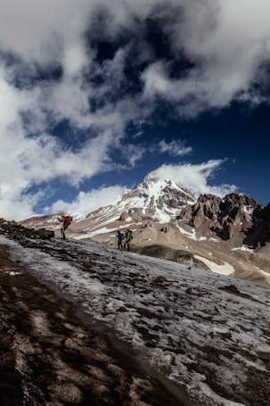 Hikers ascend a rugged, snowy mountain slope under a partially cloudy sky. A prominent, snow-capped peak dominates the background, while the foreground shows the varied textures of ice and rock. The hikers are equipped with backpacks and appear to be in mid-journey.