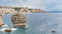 A coastal scene featuring a large, rugged rock formation emerging from the sea with layered cliffs in the background. The ocean appears calm with gentle waves, reflecting an overcast sky. A small boat with several passengers is visible on the water to the right.