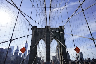 A wide-angle view of a suspension bridge with its iconic stone towers and intricate web of cables. The urban skyline is visible in the background on a clear day with a bright blue sky. Traffic cones with 'Work Zone Ahead' signs are prominently displayed.