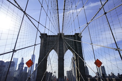 A wide-angle view of a suspension bridge with its iconic stone towers and intricate web of cables. The urban skyline is visible in the background on a clear day with a bright blue sky. Traffic cones with 'Work Zone Ahead' signs are prominently displayed.