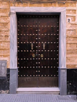 A large wooden double door with numerous metal studs and ornate handles. The door is framed by light gray marble, and the surrounding wall is made of textured, yellowish-brown bricks. There is a number '2' plaque on the right side above the door frame, and a small metallic keypad or intercom to the left.
