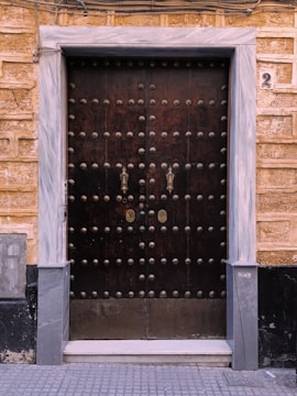 A large wooden double door with numerous metal studs and ornate handles. The door is framed by light gray marble, and the surrounding wall is made of textured, yellowish-brown bricks. There is a number '2' plaque on the right side above the door frame, and a small metallic keypad or intercom to the left.