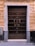 A large wooden double door with numerous metal studs and ornate handles. The door is framed by light gray marble, and the surrounding wall is made of textured, yellowish-brown bricks. There is a number '2' plaque on the right side above the door frame, and a small metallic keypad or intercom to the left.