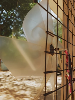 A large, translucent plastic fan is mounted on a wire grid fence. Sunlight creates intricate patterns of light and shadow on the surrounding surface. In the background, there are green trees, providing a natural setting.