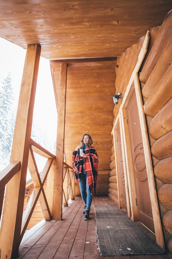 A woman in a cozy plaid shawl stands on a wooden cabin porch holding a hot drink. The cabin is constructed with logs, featuring warm wooden tones. Snow can be seen on the nearby rooftop, hinting at a wintry setting.
