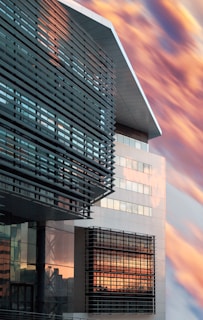 A modern building features sleek architectural lines with a mix of glass and metal panels. The windows reflect a dramatic sky filled with warm-toned clouds, creating a striking contrast between the structured design and the natural backdrop.