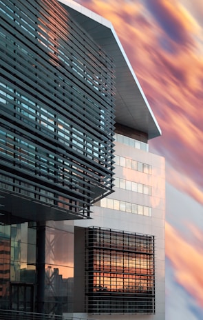 A modern building features sleek architectural lines with a mix of glass and metal panels. The windows reflect a dramatic sky filled with warm-toned clouds, creating a striking contrast between the structured design and the natural backdrop.