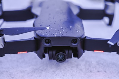 A close-up view of a black drone resting on a snowy surface. Snowflakes lightly cover its body, and the camera lens is prominently visible at the front. The drone's propellers are extended, and there are small red lights visible on its arms.