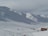 Snow-covered mountains with several people skiing on the slopes. A small building with an orange roof is visible in the foreground. Ski lifts and trails can be seen, and some groups of people are gathered near the base.