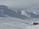 Snow-covered mountains with several people skiing on the slopes. A small building with an orange roof is visible in the foreground. Ski lifts and trails can be seen, and some groups of people are gathered near the base.