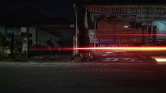 A street scene at night with a shop selling bakso and mie ayam. The shop is closed with a rolled-up bamboo curtain covering part of the entrance. Light trails from a passing vehicle create red and orange streaks across the image, suggesting movement.