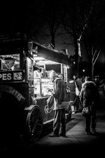 A black and white photograph features a street scene at night with a small food cart illuminated by its internal lights. A person in a coat and hat is standing in front of the cart, seemingly making a purchase. Other pedestrians walk by in the background, and the structure of the Eiffel Tower is partially visible, suggesting the location is Paris.