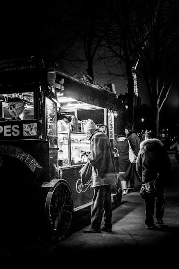 A black and white photograph features a street scene at night with a small food cart illuminated by its internal lights. A person in a coat and hat is standing in front of the cart, seemingly making a purchase. Other pedestrians walk by in the background, and the structure of the Eiffel Tower is partially visible, suggesting the location is Paris.