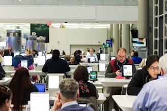 A busy airport terminal features rows of tables where people are using electronic devices like tablets and laptops. The setting appears to be a modern waiting area with bright overhead lighting and a functional design. Individuals are focused on their screens, suggesting engagement with personal tasks or communication.