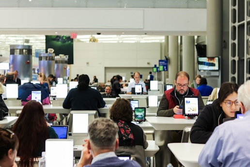 A busy airport terminal features rows of tables where people are using electronic devices like tablets and laptops. The setting appears to be a modern waiting area with bright overhead lighting and a functional design. Individuals are focused on their screens, suggesting engagement with personal tasks or communication.