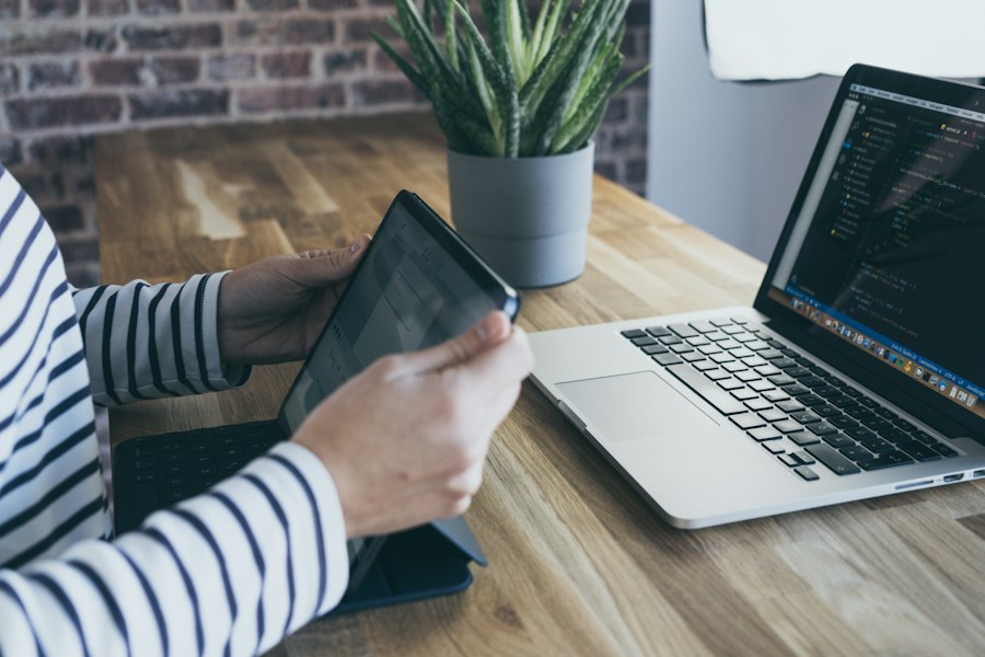 Tablet and laptop on a wooden desk beside a plant