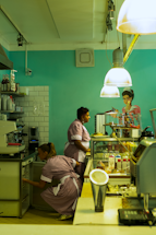 Two people in uniforms are working in a cafe or diner environment. One person is standing next to a glass display case filled with plates of food, while the other is crouched down, likely handling items inside a lower storage compartment. Overhead, large pendant lights illuminate the scene, and there's a painted mural of a woman on the wall.