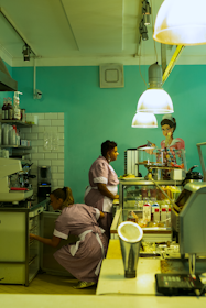Two people in uniforms are working in a cafe or diner environment. One person is standing next to a glass display case filled with plates of food, while the other is crouched down, likely handling items inside a lower storage compartment. Overhead, large pendant lights illuminate the scene, and there's a painted mural of a woman on the wall.