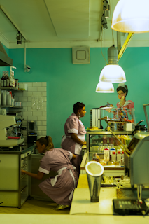 Two people in uniforms are working in a cafe or diner environment. One person is standing next to a glass display case filled with plates of food, while the other is crouched down, likely handling items inside a lower storage compartment. Overhead, large pendant lights illuminate the scene, and there's a painted mural of a woman on the wall.
