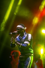 A performer wearing sunglasses and a headband is on stage, holding a microphone. Dressed in a blue tracksuit with white accents, they exude a confident stage presence. The background is vivid with colorful lighting, casting shades of yellow and red.