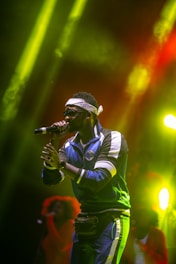 A performer wearing sunglasses and a headband is on stage, holding a microphone. Dressed in a blue tracksuit with white accents, they exude a confident stage presence. The background is vivid with colorful lighting, casting shades of yellow and red.