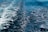 A dynamic, swirling pattern of foam on a deep blue ocean surface, created by the wake of a moving boat. The contrast between the bright white foam and the dark blue water highlights the motion and texture of the sea.