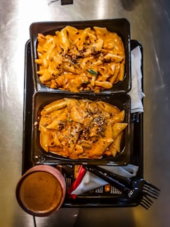 Two rectangular black trays containing creamy orange pasta topped with herbs and spices. The trays are placed on a larger black tray alongside a cup of coffee, a napkin, and two black forks. The pasta appears to be penne, covered in a rich sauce and garnished with seasoning.