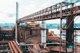 A large industrial complex featuring interconnected metal structures and pipes. The facility is expansive, situated near a body of water, with towers and walkways crisscrossing overhead. The scene appears to be a factory or refinery with visible rust and wear, indicating a long history of use.