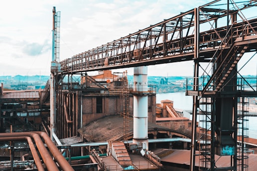 A large industrial complex featuring interconnected metal structures and pipes. The facility is expansive, situated near a body of water, with towers and walkways crisscrossing overhead. The scene appears to be a factory or refinery with visible rust and wear, indicating a long history of use.