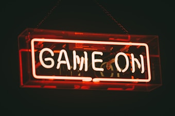 A neon sign with the words 'Game On' is enclosed in a rectangular transparent casing. The glowing letters are illuminated in a warm white color, contrasting against a dark background.