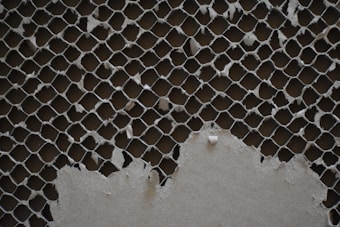 A close-up view of a piece of cardboard with a honeycomb pattern that is partially torn, revealing the texture and intricate design. The material appears worn and frayed, creating a rough and uneven surface.