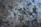 A close-up view of coarse, translucent salt crystals mixed with small, darker mineral stones. The crystals vary in size and shape, with a predominant white hue and occasional gray and brown tones.