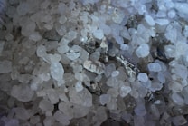 A close-up view of coarse, translucent salt crystals mixed with small, darker mineral stones. The crystals vary in size and shape, with a predominant white hue and occasional gray and brown tones.