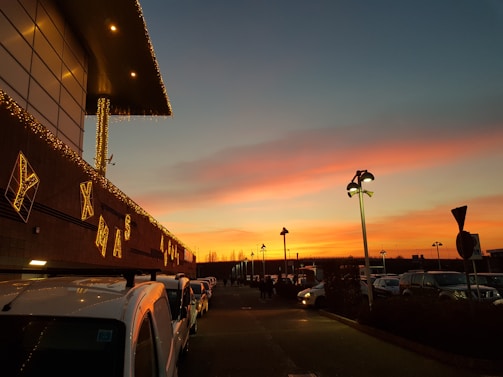 A parking lot is lined with vehicles, and a large building adorned with decorative Christmas lights is visible. The sky displays a vibrant sunset with shades of orange, pink, and blue, creating a striking backdrop. Streetlights are present, casting a warm glow, and there are various signs and poles.