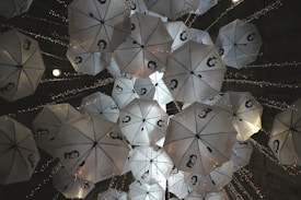 A group of white umbrellas with black and white portraits printed on them is suspended from the ceiling. They are adorned with small, twinkling string lights, creating an artistic and whimsical atmosphere. The umbrellas are arranged at different angles, providing a dynamic and intriguing visual display.