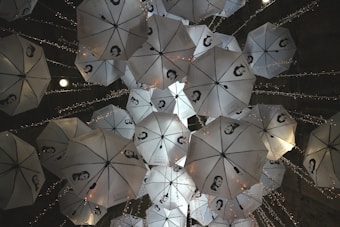 A group of white umbrellas with black and white portraits printed on them is suspended from the ceiling. They are adorned with small, twinkling string lights, creating an artistic and whimsical atmosphere. The umbrellas are arranged at different angles, providing a dynamic and intriguing visual display.